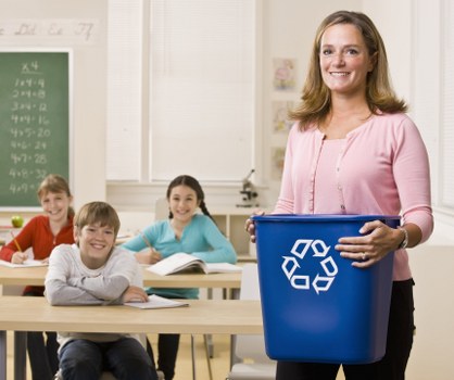 A woman in a pink top holding a blue rubbish bin with a white recycling symbol on the front stands in a classroom, smiling at the camera. Behind her, three children are seated at light wooden desks, engaged with their books and notebooks, in a setting that appears to be a school classroom with light-colored walls and a window with blinds. To the left, a green chalkboard displays mathematical calculations and numbers, indicating an educational environment. The room is well-lit with natural light from the window, and the scene suggests a focus on waste management or recycling awareness, supporting the services of a rubbish removal company like Office Clearance Hayes operating near Hayes, South West London, possibly within the HA postcode area, and reflects a professional approach to waste disposal in educational settings.
