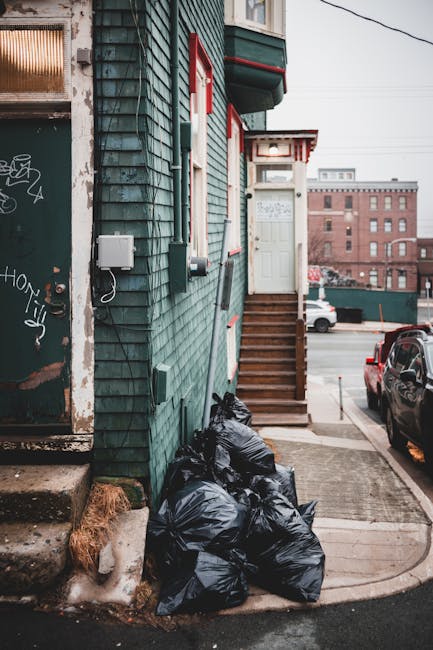 A row of black garbage bags filled with waste materials is piled against the exterior wall of a residential building in an urban setting, near the concrete steps leading to a white door on a small por