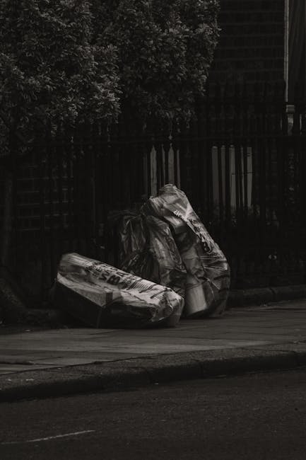 A row of black garbage bags filled with waste materials is piled against the exterior wall of a residential building in an urban setting, near the concrete steps leading to a white door on a small por
